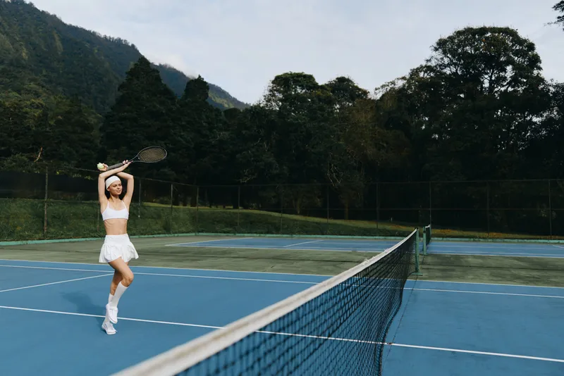 Young female tennis player in sporty outfit enjoying a game on outdoor court with mountains in the background, showcasing energy and excitement