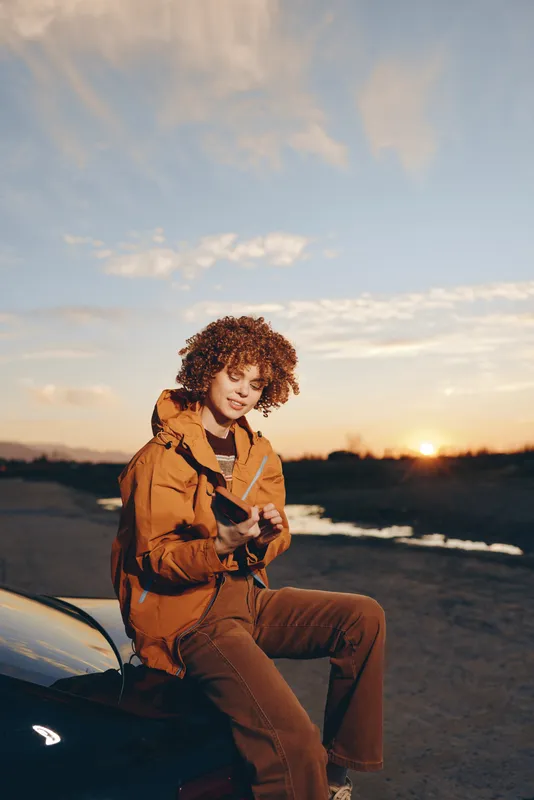 Woman lifestyle portrait outdoors at sunset beside car, curly hair woman in orange jacket, holding phone for travel, coastal evening light, relaxed adventure mood