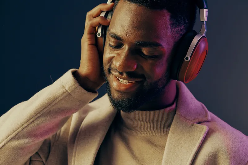 Young Black man enjoying music with headphones, wearing a stylish sweater and coat in a colorful studio setting, expressing joy and relaxation