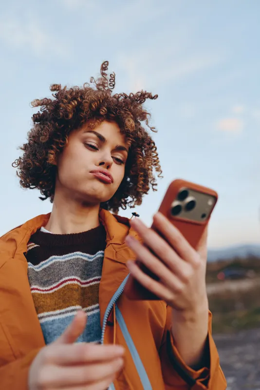 Woman lifestyle outdoors, curly hair in wind, orange jacket, holding smartphone and object, relaxed pose, sunlit face, soft countryside background, calm expression