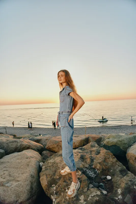 Lifestyle woman in modern boho-western denim street style standing on rocks by the ocean at sunset, captured in warm film color tones for natural mood.