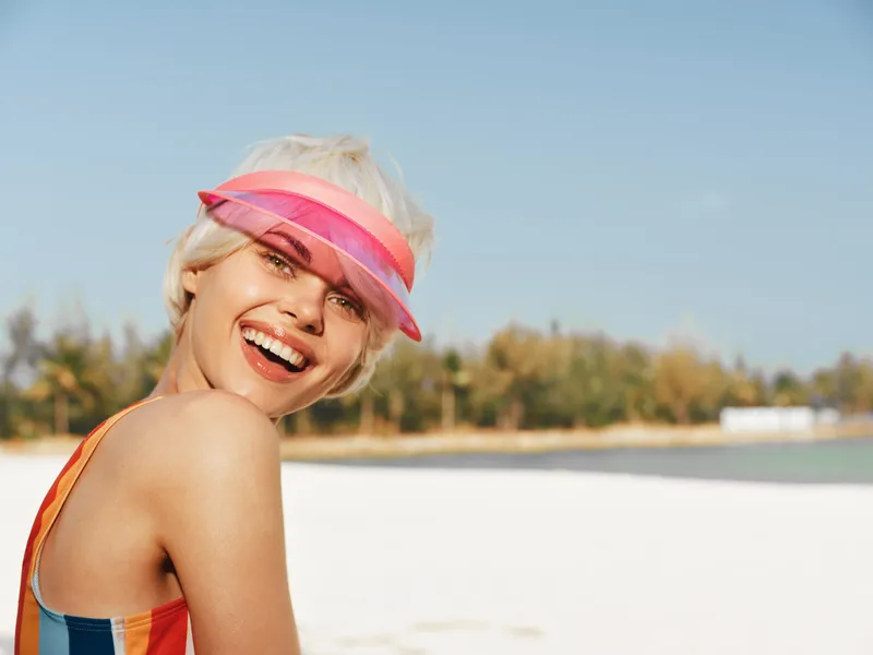 Vibrant portrait of a smiling woman on a sunny beach, wearing a colorful swimsuit and pink visor, capturing carefree summer joy against bright sea and sky.