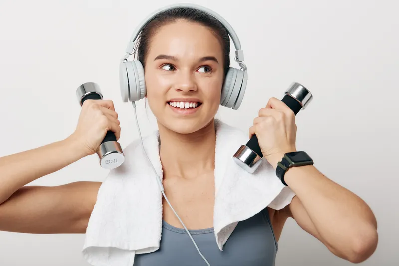 Fitness woman in a gym setting, wearing ear headphones and a towel around neck, lifting dumbbells for workout and cardio, focused on strength training and endurance.
