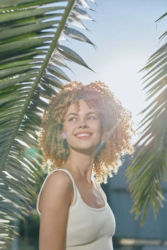 smiling woman with curly hair stands between sunlit palm leaves wearing white tank top under bright blue sky, summertime, natural light, tropical vibe