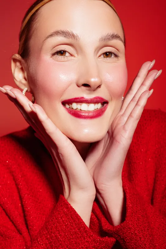 Smiling woman with glowing skin and red lipstick poses with hands near face on red background. Concept of beauty, skincare, happiness, and vibrant makeup in studio lighting.
