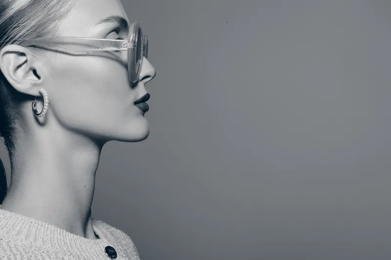 Profile of a young white woman with fair skin wearing transparent glasses and hoop earrings on a grey background. Concept of fashion, style, and modern beauty.