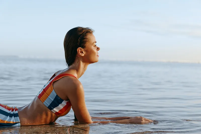 Young woman in striped bikini rests at the water edge, gazing toward the horizon as gentle waves lap her torso in warm, sunlit calm.