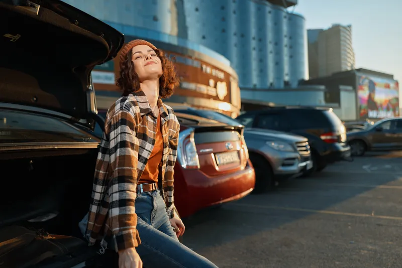 Woman leaning on car trunk in parking lot during golden hour, candid lifestyle portrait showing authenticity and relaxed joy, urban fashion, mindful living and emotional storytelling mood.