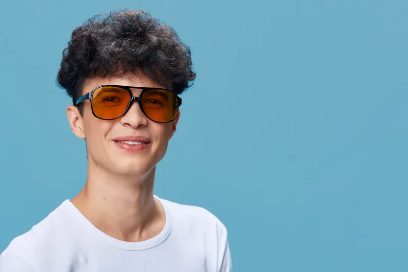 smiling young man with curly hair wearing orange sunglasses and white t-shirt on blue background, casual style, positive expression, studio portrait