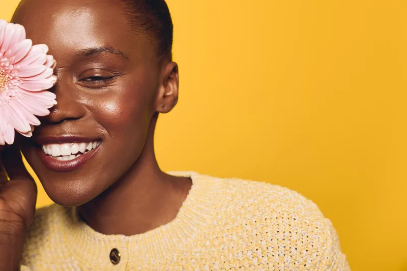 Happy African woman with dark skin smiles and holds a pink flower near her face on yellow background. Concept of natural beauty, skincare, wellness, and positive mood.