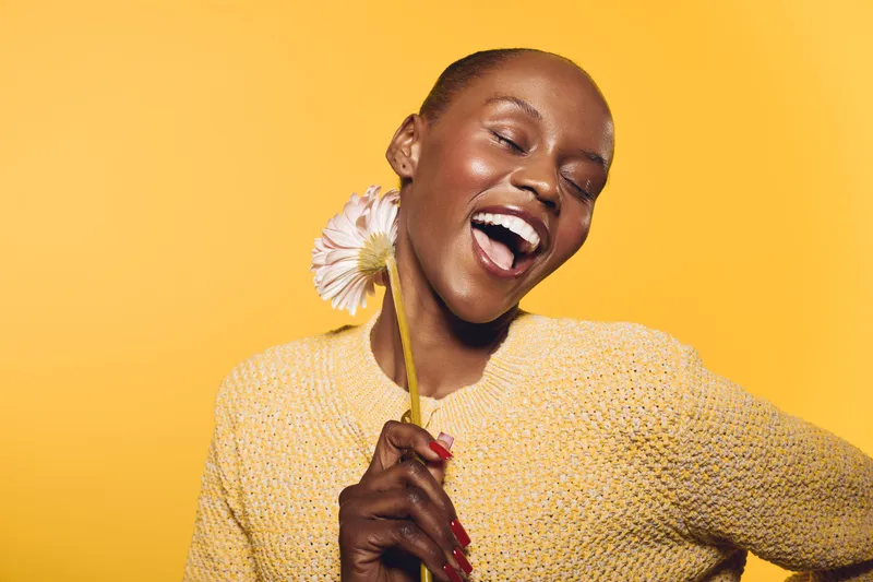 Joyful African woman with dark skin laughing and holding a flower near her face. She wears a yellow sweater against a bright yellow background. Concept of happiness and natural beauty.