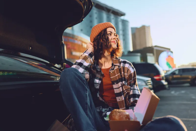 Woman sits at car trunk in a parking lot, urban sunset scene with candid lifestyle vibe, enjoying takeout. Golden hour glow and authenticity highlight mindful living and emotional storytelling.