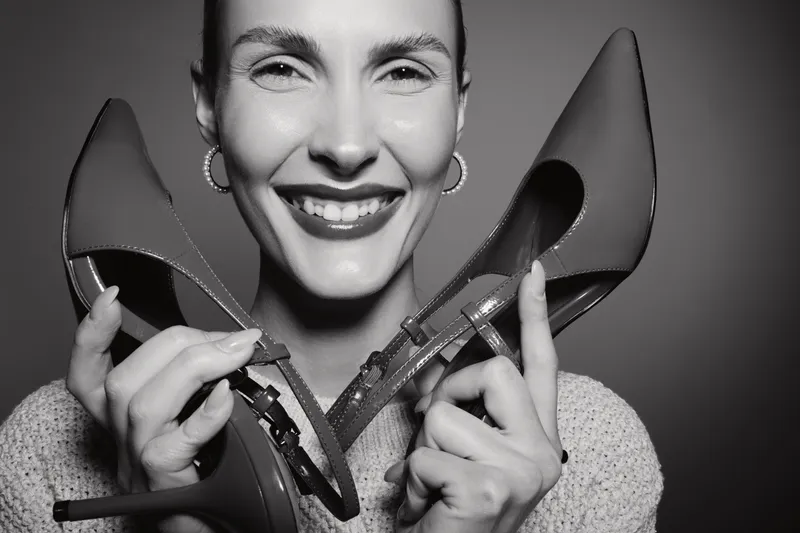 Happy white woman with fair skin smiling and holding high heel shoes close to face in studio. Concept of fashion, footwear, and female lifestyle.
