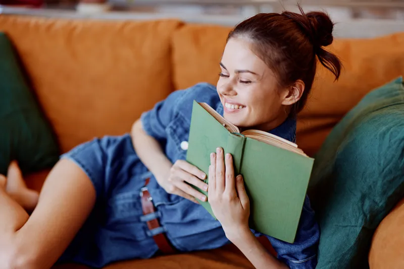 Счастливая женщина сидит на диване, читает книгу и улыбается в камеру в уютной гостиной