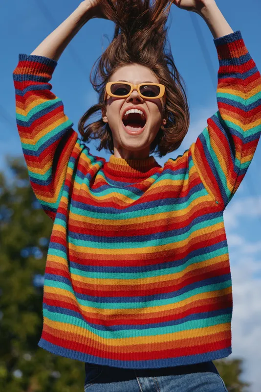 Woman in rainbow sweater and sunglasses smiling joyfully, portrait of a young woman outdoors with hands raised, colorful knit fashion and carefree summer vibe.