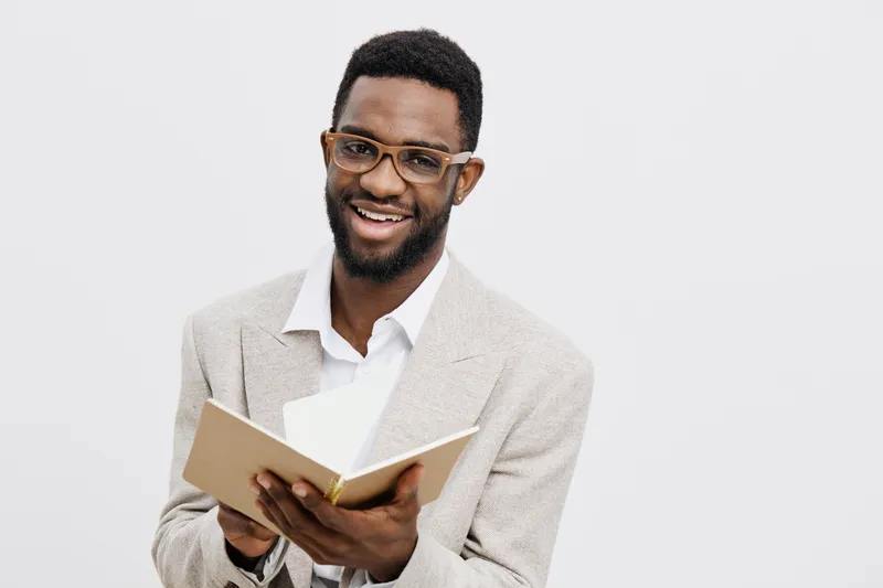 Smiling man with positive expression reading a notebook in a studio setting, dressed in a light-colored blazer and white shirt, wearing glasses, creating a professional and friendly atmosphere. People
