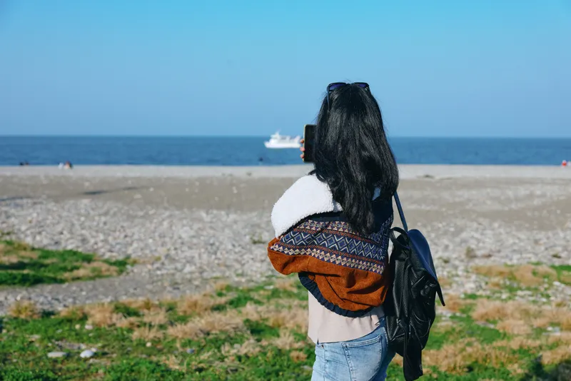 Woman with backpack on a sunny beach facing the sea, wrapped in a scarf and sweater, exploring the coast with a calm travel mood and seaside outdoor leisure