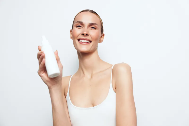 Smiling woman in white tank top holds white bottle skincare product looks at camera isolated on white background with positive expression and natural makeup.