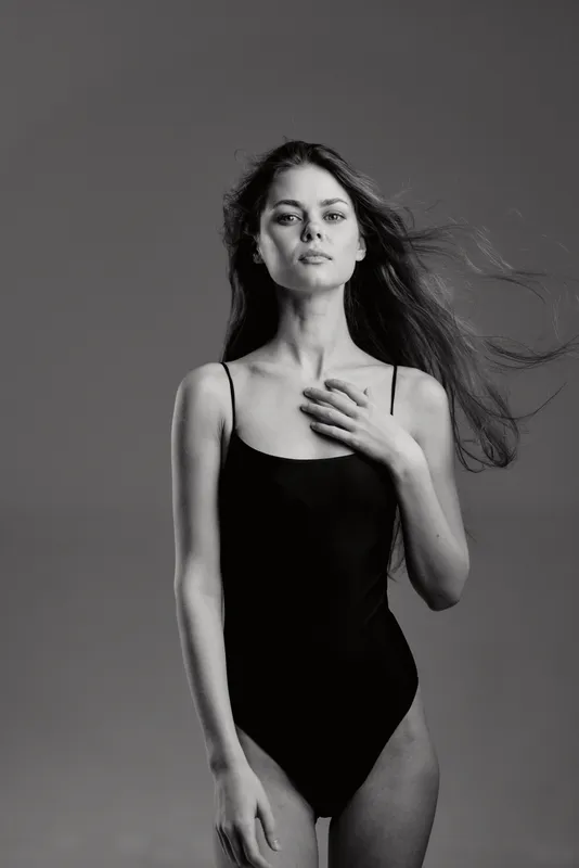 Black and white portrait of a young woman in a black swimsuit with flowing hair, expressing calm and confidence in a studio setting.