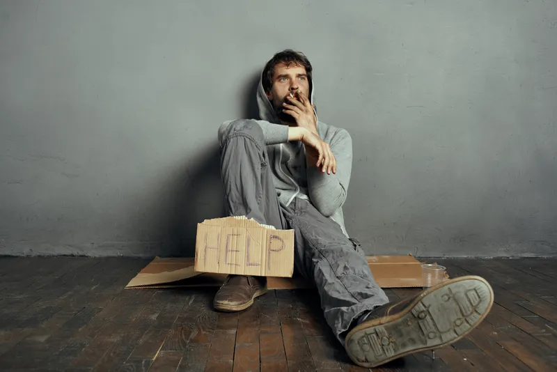 Бородатый бомж сидит на полу, держа табличку «Help»