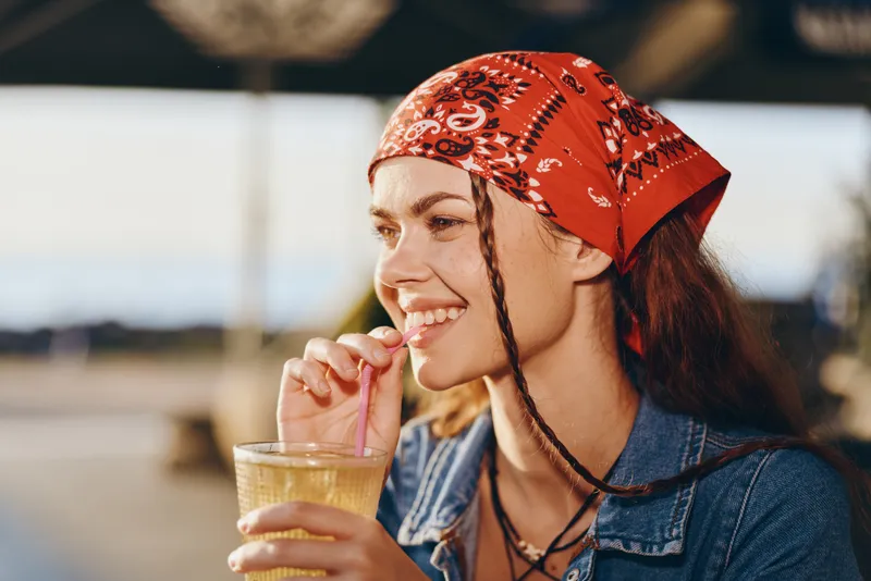 Lifestyle woman in modern boho-western denim street style enjoying a drink, smiling joyfully with film color warmth and natural outdoor background.
