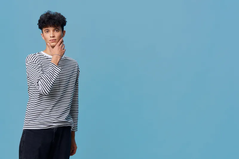 Young man in striped shirt posing thoughtfully against plain blue background, casual style, curly hair, minimalistic composition, youth lifestyle concept.