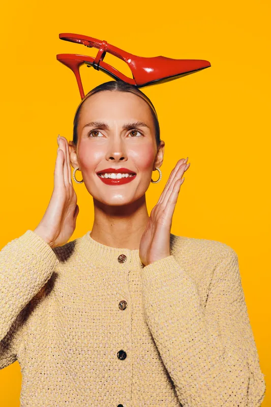 Smiling woman with red high heel shoe on head poses against bright yellow background. Fashion concept with playful mood and creative styling in studio portrait of happy model.