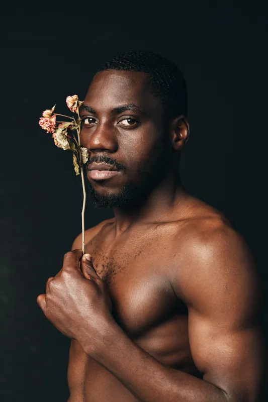 African man with dark skin holding dried flower close to face in creative conceptual visual style. Portrait of shirtless male model with thoughtful expression on black background.