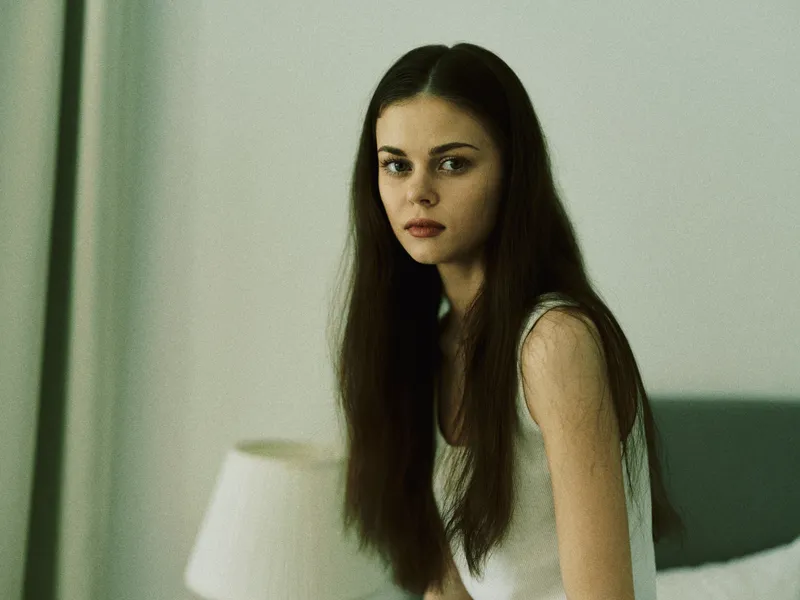 Serious young woman with long hair sitting indoors in soft natural light, wearing a white sleeveless top, with a calm and thoughtful expression.