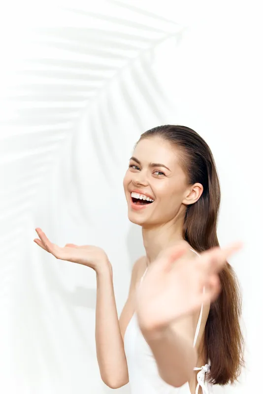 Радостная молодая женщина в белом платье излучает счастье и позитив, мягкие тени от пальмовых листьев падают на яркий фон