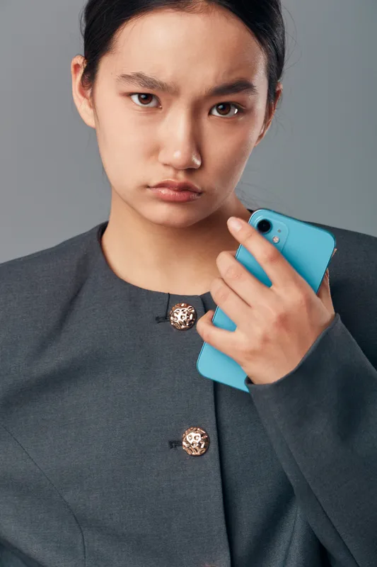 Young woman holding blue smartphone with serious expression in studio portrait. Female wearing dark jacket looking worried or concerned with phone in hand against gray background.