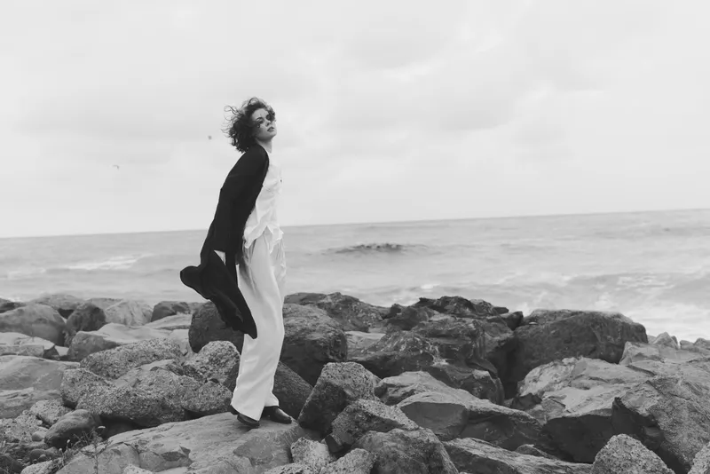 Black and white scene of a woman standing on jagged rocks by the sea, wind in hair, calm water in distance, moody coastal atmosphere, balance between nature and human form, contemplative moment on a