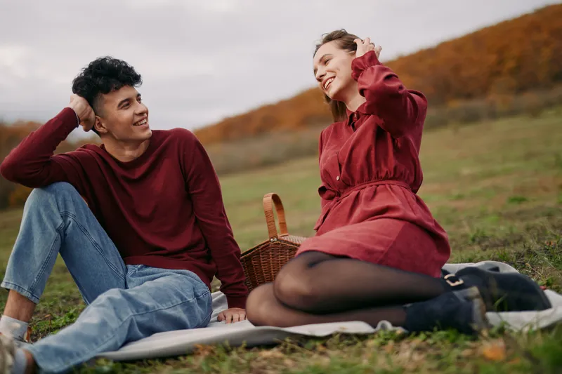 A candid outdoor portrait of a young couple sharing smiles on a picnic blanket, capturing genuine connection and relaxed authenticity with natural expressions and warm eye contact.