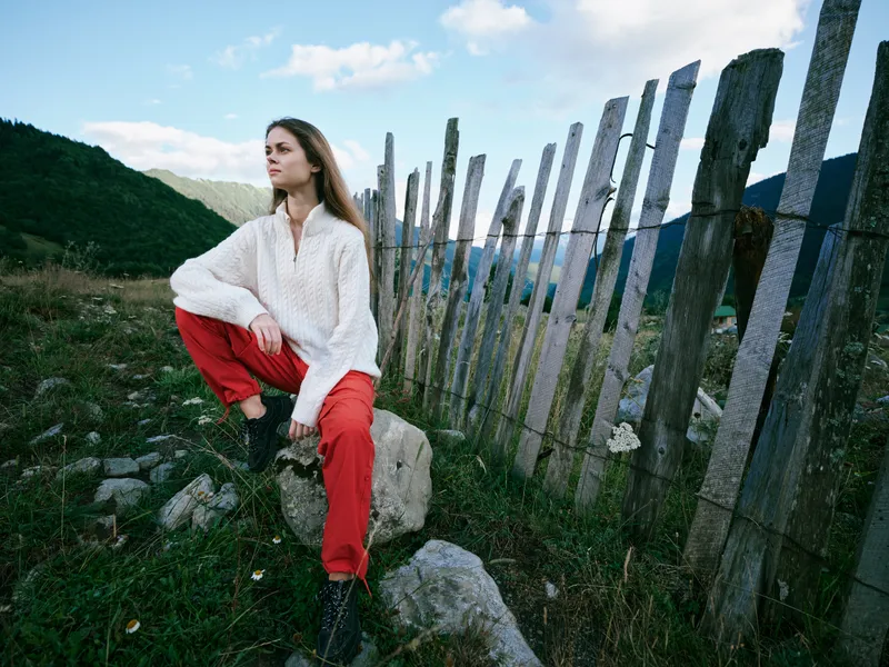 A woman sits perched on rocks beside a weathered fence in a countryside landscape, wearing a white sweater and red pants, capturing a serene, rustic moment outdoors.