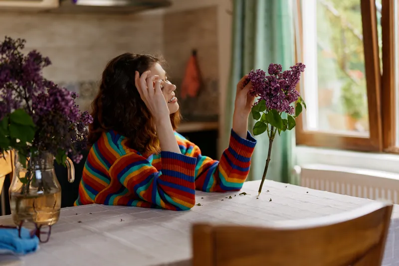 A woman in a bright striped sweater sits at a sunlit kitchen table, resting her head on her hands while admiring lilac flowers by a large window, capturing cozy home moments.