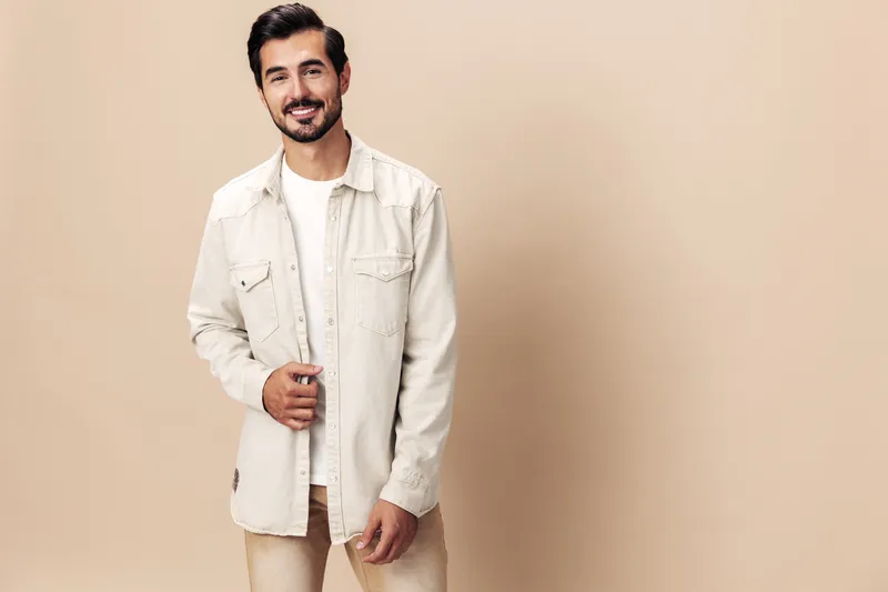 Стильный мужчина с улыбкой, модель позирует на бежевом фоне в белой футболке, смотрит в камеру, модный стиль одежды