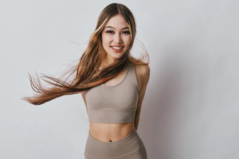 Young woman with long hair wearing sportswear smiling and posing isolated on light background. Athletic female model showing fitness lifestyle and natural beauty in studio.