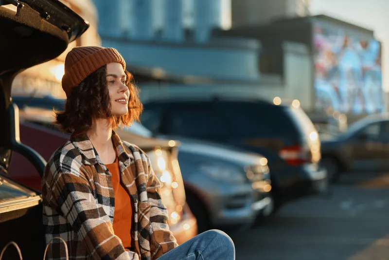 woman portrait candid golden urban parking sunlight beanie relaxed lifestyle Young woman sits on car trunk in parking lot at golden hour glow, authenticity and mindful living captured with emotional