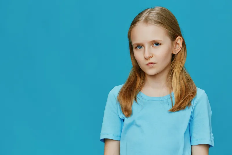 A serious young girl with long straight hair wearing a light blue shirt against a vibrant blue background, conveying a sense of introspection and calmness