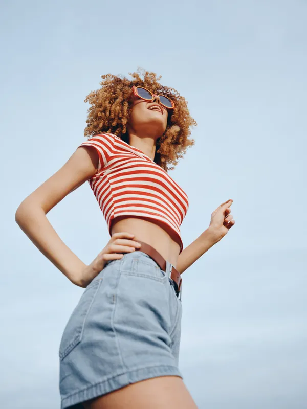 A young woman stands proudly outdoors, wearing a red and white striped crop top, high waist denim shorts, sunglasses, and a confident pose against a bright blue sky.