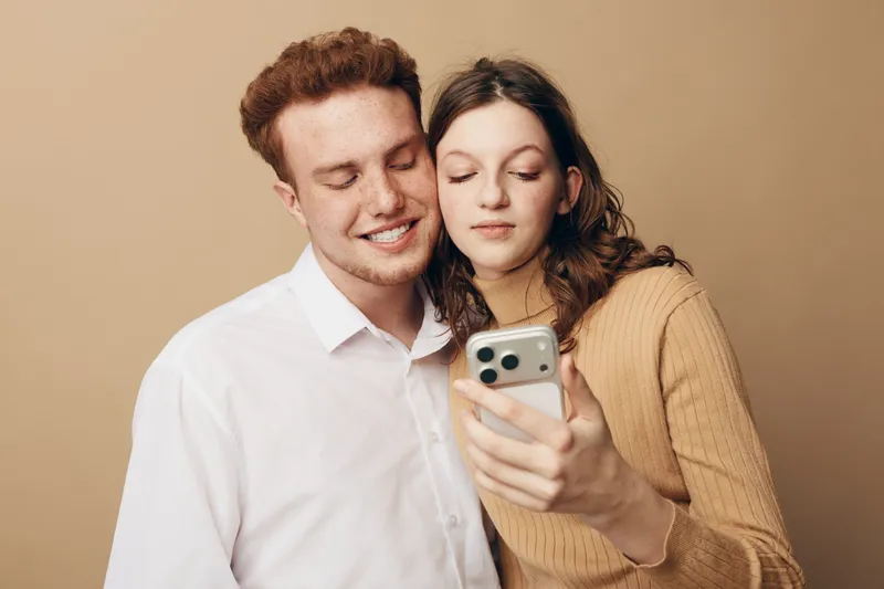 Young couple with closed eyes smiling while looking at smartphone together. They stand close in casual clothes against beige background. Concept of love, technology, and relationship.
