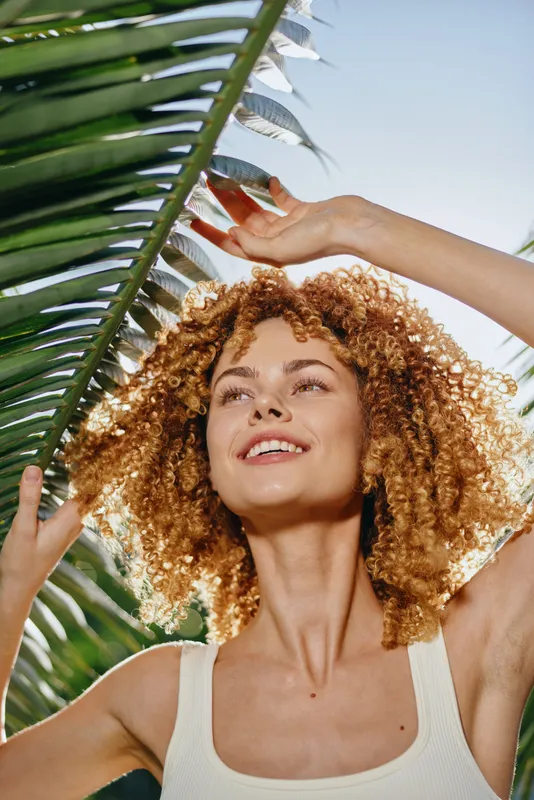smiling woman with curly hair enjoying sunny tropical day near green palm leaves, natural light, warm mood, summer outdoor portrait
