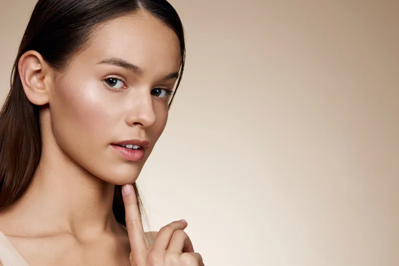 Elegant young woman with a natural makeup look, posing against a soft beige background, expressing a calm and thoughtful emotion in a seasonal beauty concept