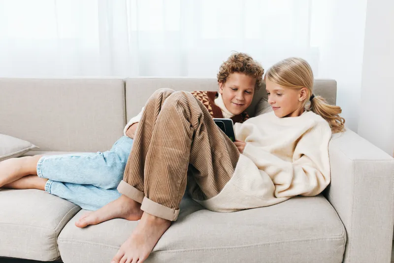 Authentic scene of siblings relaxing at home, enjoying a tablet together, showcasing a playful study vibe and casual style in a cozy living room bathed in bright light and everyday details of family