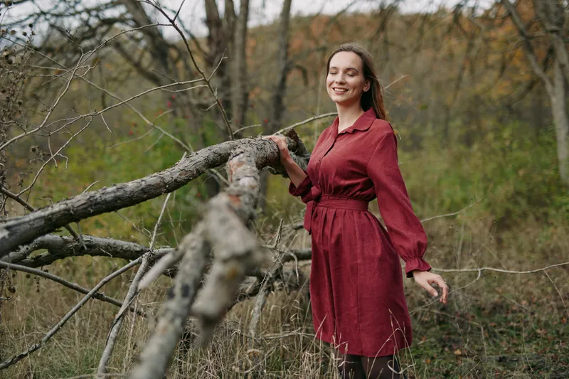 An authentic scene captures a smiling woman in a burgundy dress beside a fallen branch, presenting genuine mood and natural light in a quiet rural setting.