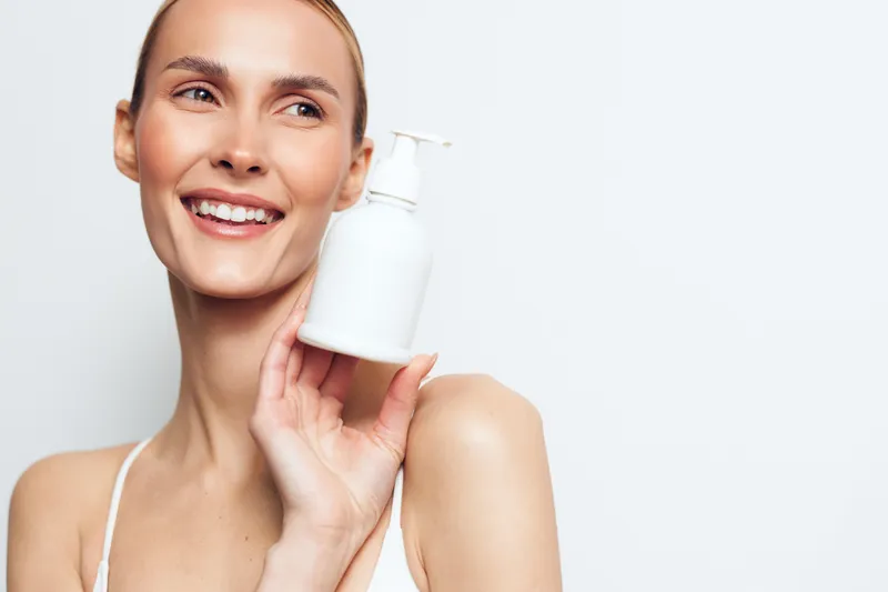 Smiling young woman with blonde hair holds a white lotion bottle near her face on a plain light background conveying skincare and self-care concepts.