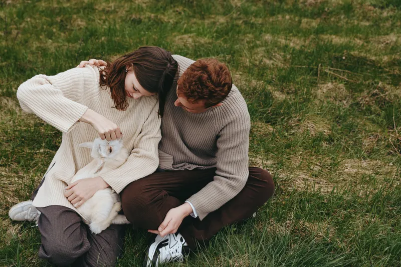 Couple outdoors on grass with a small white dog, sharing a tender moment. Cozy sweaters, natural light, intimate scene emphasizing companionship, nature, and simple everyday joy.