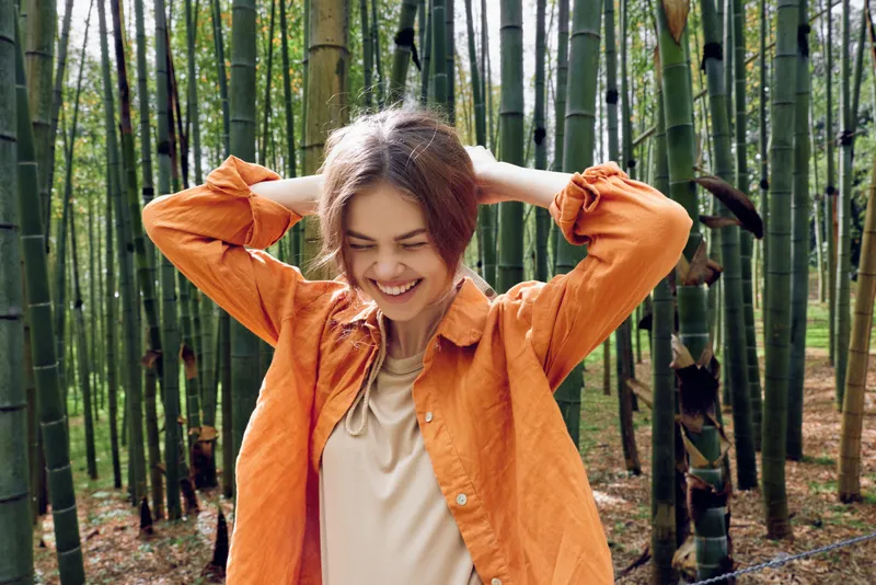 Woman smile in orange jacket standing in bamboo forest, portrait outdoors in nature. Happy female traveler with casual style, relaxed expression and candid pose among green bamboo stalks and woods