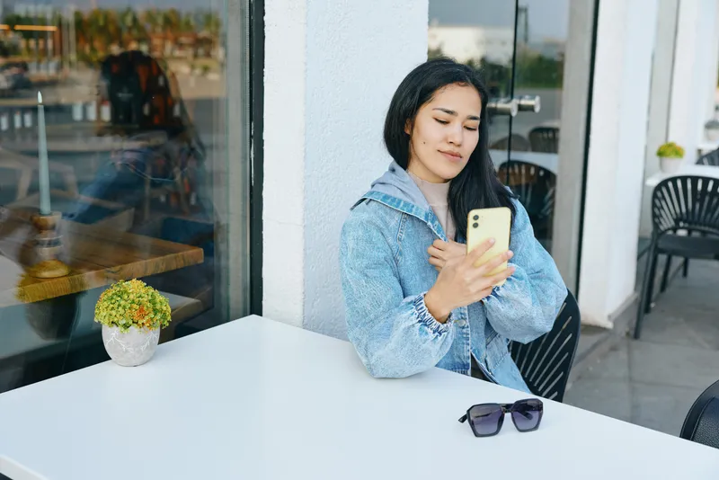 Woman sits at an outdoor cafe table, looking at smartphone while wearing a denim jacket. Sunglasses rest on table, potted plant nearby, glass door backdrop, candid urban moment.