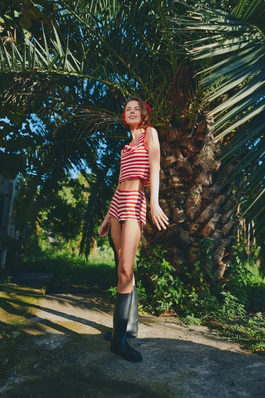 A confident young woman in a red and white striped bikini stands beside a palm tree on a sunny outdoor path, wearing tall black boots and smiling at the camera.